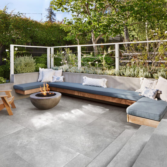 Terras met een lange bank met blauwgrijze kussens, een ronde vuurschaal en een grijze betonlook tegelvloer
