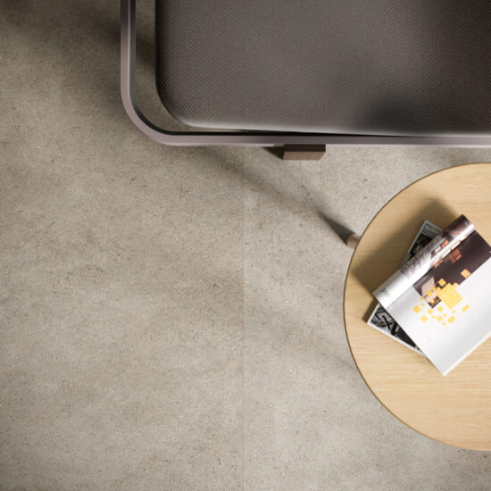 Bovenaanzicht van een lichtgrijze betonlookvloer met één dunne voeglijn, met de rand van een donkere fauteuil en een rond eiken bijzettafeltje